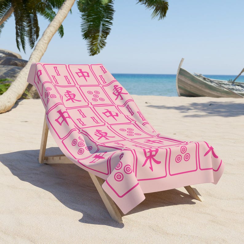 May include: A pink beach towel with a pink mahjong tile pattern. The towel is draped over a wooden beach chair. The towel has Chinese characters and circles on it.