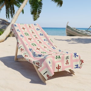 May include: A pink beach chair with a white and pink beach towel. The towel has a pattern of Chinese mahjong tiles with black, red, blue, and green characters. The beach chair is on a white sandy beach with palm trees and a blue ocean in the background.