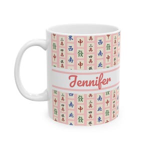 May include: White ceramic mug with a pink background featuring a pattern of Mahjong tiles. The mug has the name "Jennifer" in a cursive font. The handle is white.