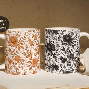 May include: Two ceramic mugs with floral patterns. One mug features orange flowers and leaves on a white background, the other has black flowers and leaves. Both mugs have the text "f*ck Trump" subtly incorporated into the design.