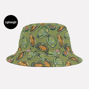 May include: A lightweight bucket hat with a repeating pattern of green and gold oyster illustrations on a muted green background. The hat has a rounded crown and a downward-sloping brim. The word "Lightweight" is in a black circle.