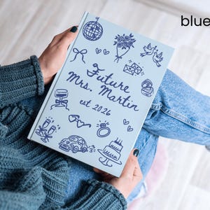 May include: A light blue journal with the text "Future Mrs. Martin est 2026" and various doodle illustrations in dark blue. The journal is held by a person wearing a blue sweater and jeans.