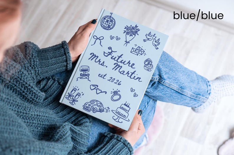 May include: A light blue journal with the text "Future Mrs. Martin est 2026" and various doodle illustrations in dark blue. The journal is held by a person wearing a blue sweater and jeans.