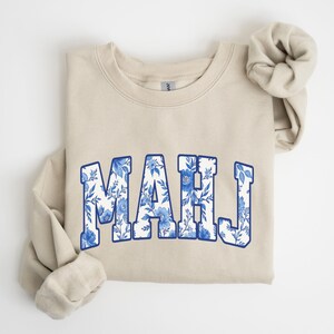 May include: A sand-colored sweatshirt with the word "MAHJ" in a blue floral design. The letters are outlined in blue and filled with a white and blue floral pattern. The sleeves are folded.