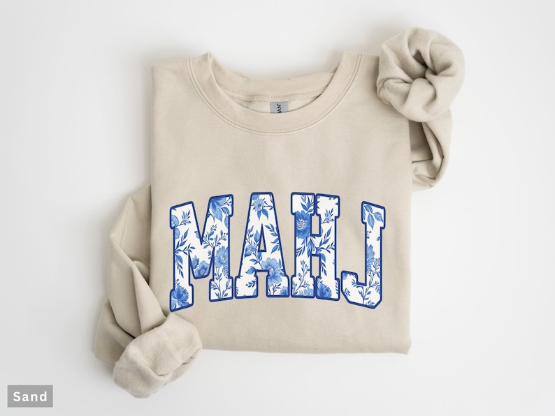 May include: A sand-colored sweatshirt with the word "MAHJ" in a blue floral design. The letters are outlined in blue and filled with a white and blue floral pattern. The sleeves are folded.