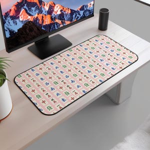 May include: A rectangular desk mat with a pink background and a repeating pattern of Mahjong tiles in red, green, blue, and black. The mat has a black border and is designed to protect a desk surface.