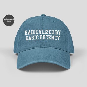 May include: A blue denim baseball cap with the text "RADICALIZED BY BASIC DECENCY" in white. The words "embroidered denim" are in a circle on the left side. The cap is a muted blue colour, suitable for casual wear.