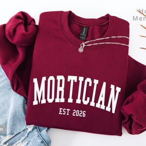 May include: A maroon sweatshirt with the word "MORTICIAN" in white arched lettering and "EST 2026" below. A silver necklace with a circular pendant rests on the sweatshirt. The sweatshirt is folded on top of a pair of blue jeans.