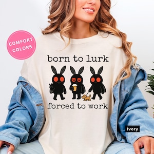 May include: Ivory-colored t-shirt with the text "born to lurk forced to work" and three cartoon rabbit-like creatures. The creatures are black with red eyes, and one holds a coffee cup, another a peace sign, and the last one walks a dog.