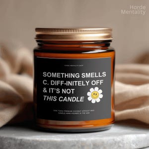 May include: A scented candle in a glass jar with a gold lid and a black label. The label features the text "SOMETHING SMELLS C. DIFF-INITELY OFF & IT'S NOT THIS CANDLE" and a smiling daisy. The candle is made with coconut apricot wax.