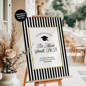 May include: A graduation sign with a black and white striped border. The sign features the text "Congratulations" and the name "Dr. Alicia Smith, Ph.D." with details about her degree and university. The sign is displayed on a wooden easel.