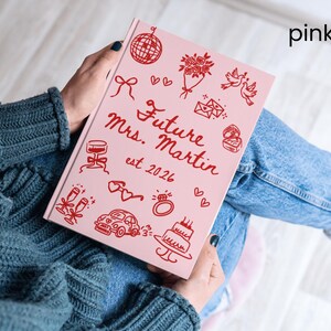 May include: A pink journal with red illustrations and text. The cover reads "Future Mrs. Martin est 2026" with drawings of a disco ball, flowers, doves, and a cake. The journal is held on a person's lap.