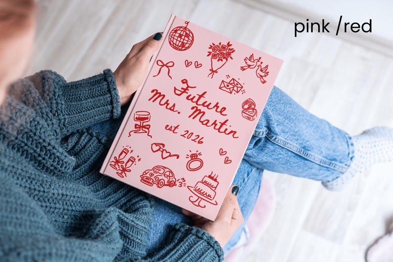 May include: A pink journal with red illustrations and text. The cover reads "Future Mrs. Martin est 2026" with drawings of a disco ball, flowers, doves, and a cake. The journal is held on a person's lap.
