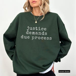 May include: A forest green sweatshirt featuring the white text "justice demands due process". The sweatshirt has a crew neck and long sleeves. The text is centrally placed on the front of the garment.