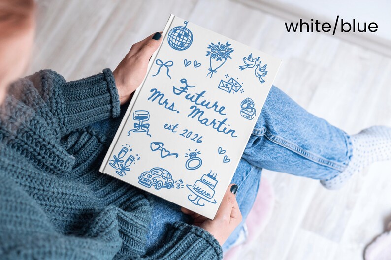 May include: A white notebook with blue illustrations and text. The cover features drawings of a disco ball, flowers, doves, and a cake. The text reads "Future Mrs. Martin est 2026". The notebook is held in a person's lap.