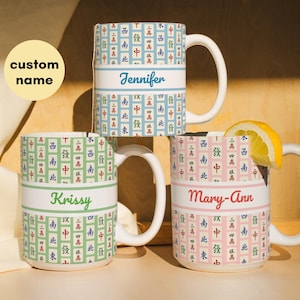 May include: Three white ceramic mugs with a Mahjong tile pattern in blue, green, and pink. Each mug has a white band with a custom name: Jennifer, Krissy, and Mary-Ann. A lemon slice rests on one mug.