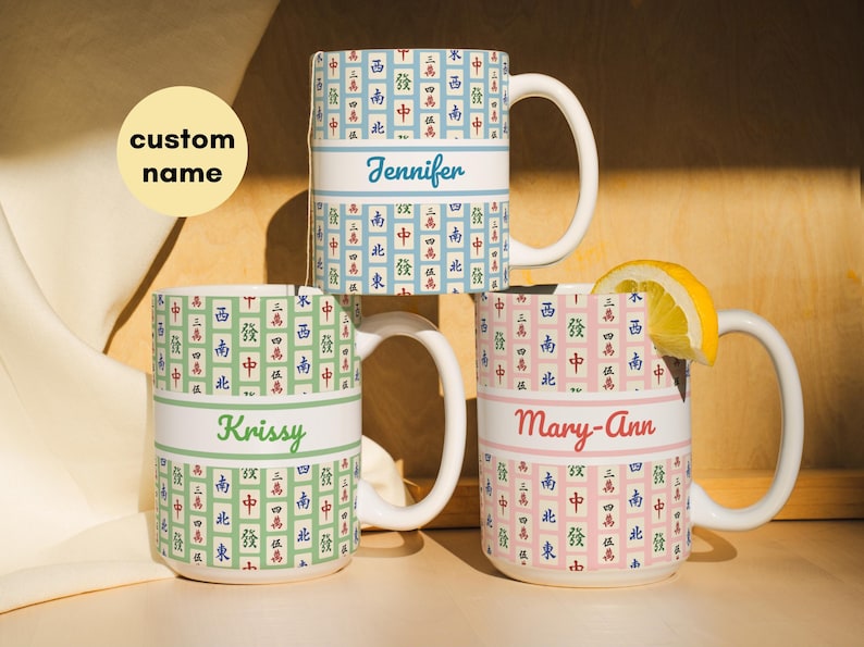 May include: Three white ceramic mugs with a Mahjong tile pattern in blue, green, and pink. Each mug has a white band with a custom name: Jennifer, Krissy, and Mary-Ann. A lemon slice rests on one mug.