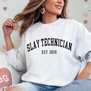 May include: White crewneck sweatshirt with the text "SLAY TECHNICIAN EST 2026" in black, arched lettering. The sweatshirt is a long-sleeved, casual top. The background includes a decorative heart-shaped pillow and a floral arrangement.