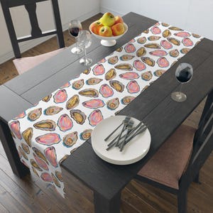 May include: A white table runner with a repeating pattern of pink and gold oyster illustrations. The runner is placed on a dark wooden table set for a meal, with plates, silverware, and wine glasses.