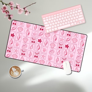 May include: A pink desk mat with a black border featuring a repeating pattern of seashells, cherries, and starfish. A pink keyboard and white mouse are on the mat. A cup of coffee and pink flowers are also in the image.