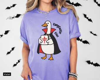May include: Violet-colored t-shirt featuring a cartoon duck dressed as a vampire, holding a Mahjong tile. The text "fangtastic tiles" is printed on the shirt. The shirt has rolled-up sleeves.