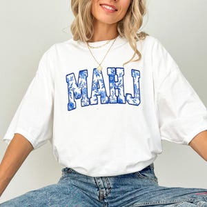 May include: White t-shirt with the word "MAHJ" in blue floral print lettering. The shirt has a relaxed fit and short sleeves. The design features a blue floral pattern within the letters, creating a stylish and eye-catching look.