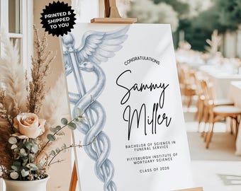Può includere: Un cartello di laurea bianco su un cavalletto con la scritta "Congratulazioni Sammy Miller" e un simbolo del Caduceo. Il cartello include anche il testo "Laurea in Scienze del Servizio Funebre".