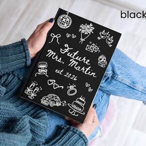 May include: A black notebook with white illustrations and text. The cover reads "Future Mrs. Martin est 2026" with drawings of wedding-related icons. The notebook is held on a person's lap, who is wearing a blue sweater and jeans.