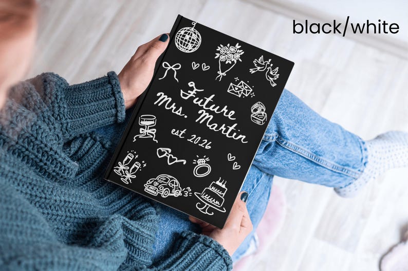 May include: A black notebook with white illustrations and text. The cover reads "Future Mrs. Martin est 2026" with drawings of wedding-related icons. The notebook is held on a person's lap, who is wearing a blue sweater and jeans.