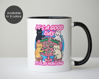 Peut inclure: Mug en céramique blanche avec une anse et un bord noirs. L'illustration représente quatre chats jouant au mahjong autour d'une table rose. Le texte sur la tasse dit "IT'S A GOOD DAY TO PLAY MAHJONG".