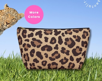 May include: A tan cosmetic bag with a leopard print pattern. The bag has a black zipper and a black trim. The words "More Colors" are in a pink circle. A leopard is in the upper left corner. The bag is on a bed of green grass.