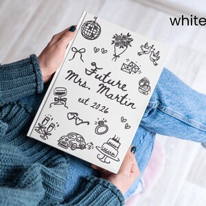 May include: A white notebook with black line art illustrations and the text "Future Mrs. Martin est. 2026". The notebook features drawings of a disco ball, flowers, doves, and a wedding cake. The notebook is held on a person's lap.