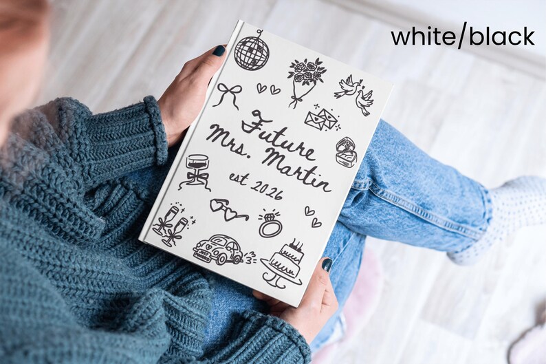 May include: A white notebook with black line art illustrations and the text "Future Mrs. Martin est. 2026". The notebook features drawings of a disco ball, flowers, doves, and a wedding cake. The notebook is held on a person's lap.