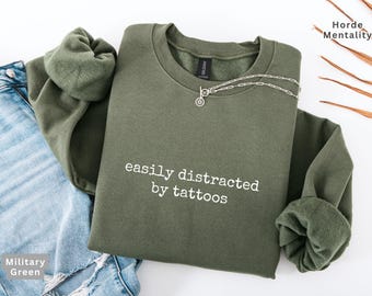 May include: An olive green sweatshirt with the text "easily distracted by tattoos" in white. A silver necklace with a pendant is on the sweatshirt. The sweatshirt is paired with blue denim jeans.