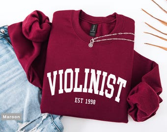 May include: A maroon sweatshirt displaying the word "VIOLINIST" in white, with "EST 1998" underneath. A silver necklace with a circular pendant is draped across the top. Light blue denim jeans are visible on the left.