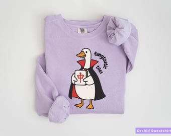 May include: Orchid-colored sweatshirt featuring a cartoon duck dressed as a vampire, holding a tile. The duck wears a black cape with a red collar. The text "fangtastic tiles" is printed on the sweatshirt.