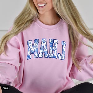 May include: A light pink sweatshirt with the word "MAHJ" in large, blue floral letters across the chest. The sweatshirt is a crew neck style and appears to be made of a soft material. The model is smiling and wearing black pants.