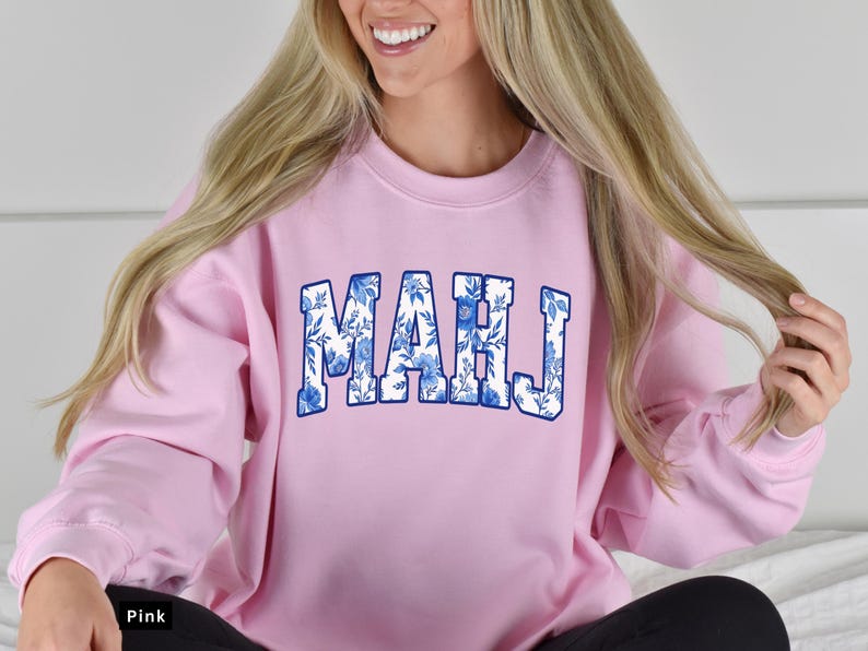May include: A light pink sweatshirt with the word "MAHJ" in large, blue floral letters across the chest. The sweatshirt is a crew neck style and appears to be made of a soft material. The model is smiling and wearing black pants.