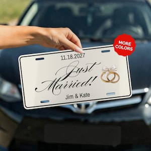Peut inclure: Plaque d'immatriculation crème avec l'inscription "Just Married!" en écriture élégante, la date "11.18.2027" et les noms "Jim & Kate". Le design comprend des alliances dorées et un nœud.