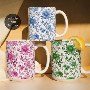 May include: Three ceramic mugs with floral patterns in blue, pink, and green. Each mug has a white handle and the words "Subtle F*ck Trump" are printed on the mugs. A lemon slice rests on the green mug.