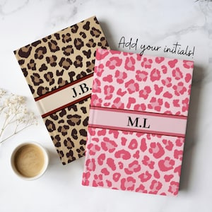 May include: Two personalized notebooks with leopard print covers. One is tan and brown, the other is pink. Each has a band with initials: "J.B." and "M.L." The text "Add your initials!" is also visible.
