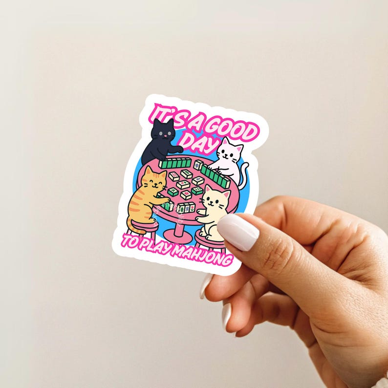 May include: A sticker featuring a cartoon illustration of four cats playing mahjong around a pink table. The text "IT'S A GOOD DAY TO PLAY MAHJONG" is written in pink and white. The cats are black, orange, and white.