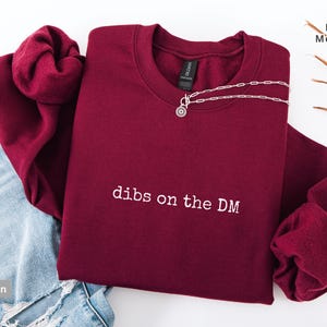 Op de afbeelding: Bordeaux sweatshirt met de tekst "dibs on the DM" in het wit. Een zilveren ketting met een ronde hanger is over de voorkant gedrapeerd. Het sweatshirt ligt naast een blauwe jeans.