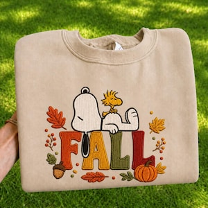 Könnte beinhalten: Ein beigefarbenes Sweatshirt mit Rundhalsausschnitt und einer Stickerei von Snoopy und Woodstock, die auf dem Wort FALL liegen. Das Design enthält herbstliche Elemente wie orangefarbene und gelbe Blätter, eine braune Eichel und einen Kürbis.