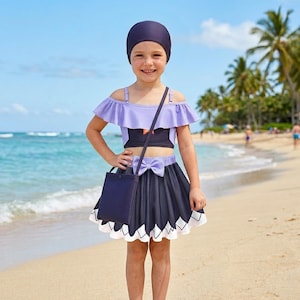 Puede incluir: Un conjunto de traje de baño de dos piezas para niños, con un top lavanda con hombros descubiertos, una falda azul oscuro con borde festoneado blanco y un gorro de baño a juego. También se incluye un bolso de mano azul oscuro. Escena de playa.