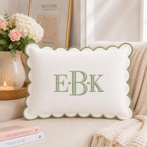 Op de afbeelding: Een rechthoekig wit sierkussen met een geschulpte rand afgewerkt in saliegroen. Het midden van het kussen toont de geborduurde monogramletters E, B en K in saliegroene draad, gescheiden door kleine saliegroene stippen.
