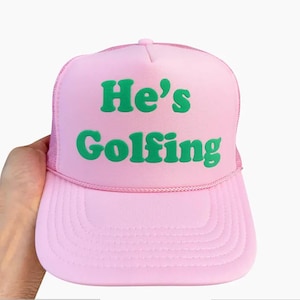 May include: A pink trucker hat with a neon green puff print that says "He's Golfing".