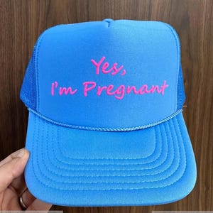 May include: A blue trucker hat with a mesh back and a curved brim. The hat features the text "Yes, I'm Pregnant" in neon pink. The hat is a vibrant blue color.