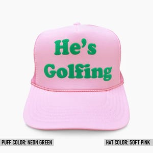 May include: A pink trucker hat with a neon green puff print that says "He's Golfing".