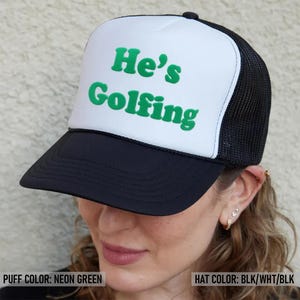 May include: A black and white trucker hat with a neon green puff print that says "He's Golfing".